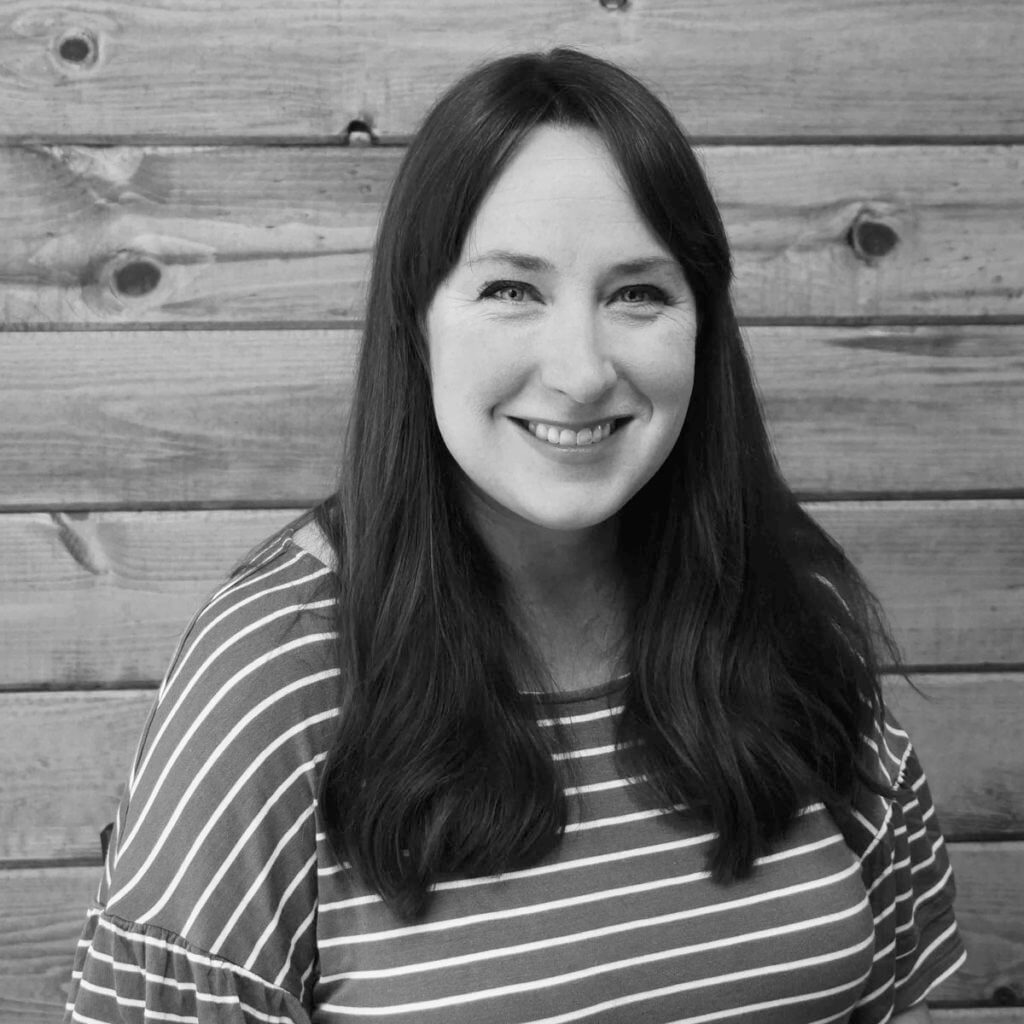 black and white portrait of a smiling woman with long dark hair, wearing a striped top, against a wooden plank background