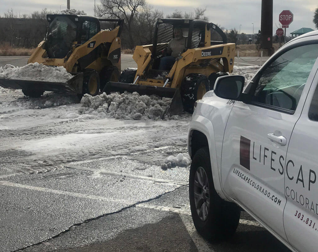 lifescape Colorado truck parked next to two yellow skid steers (CAT and DEERE) clearing snow from a parking lot