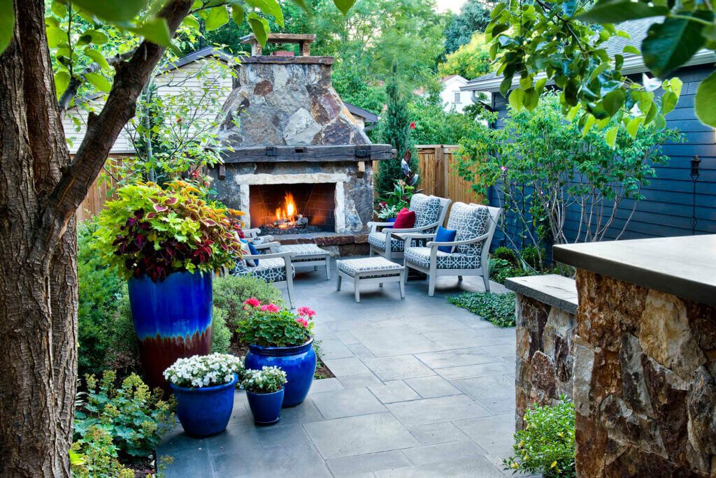 backyard patio with a large stone outdoor fireplace, stylish outdoor seating, and large blue planters with colorful foliage and flowers