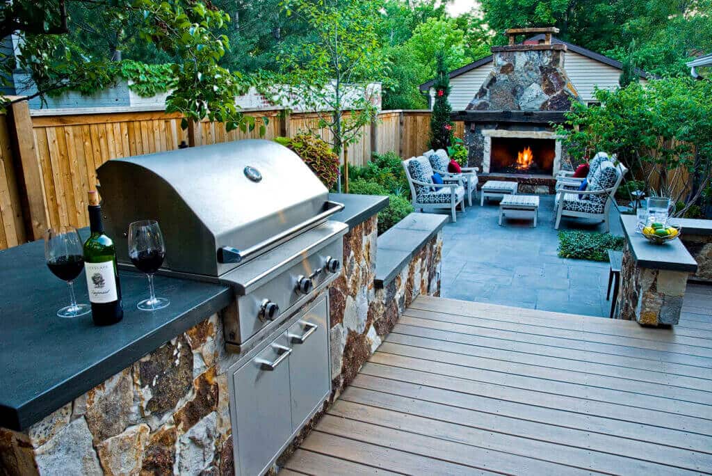 outdoor kitchen area with a built-in stainless steel grill, stone veneer base, and dark countertop, with a stone fireplace and seating area in the background