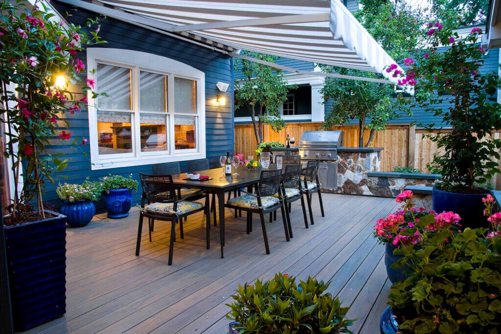 small backyard deck with outdoor dining set, striped awning, and potted flowers next to a blue house