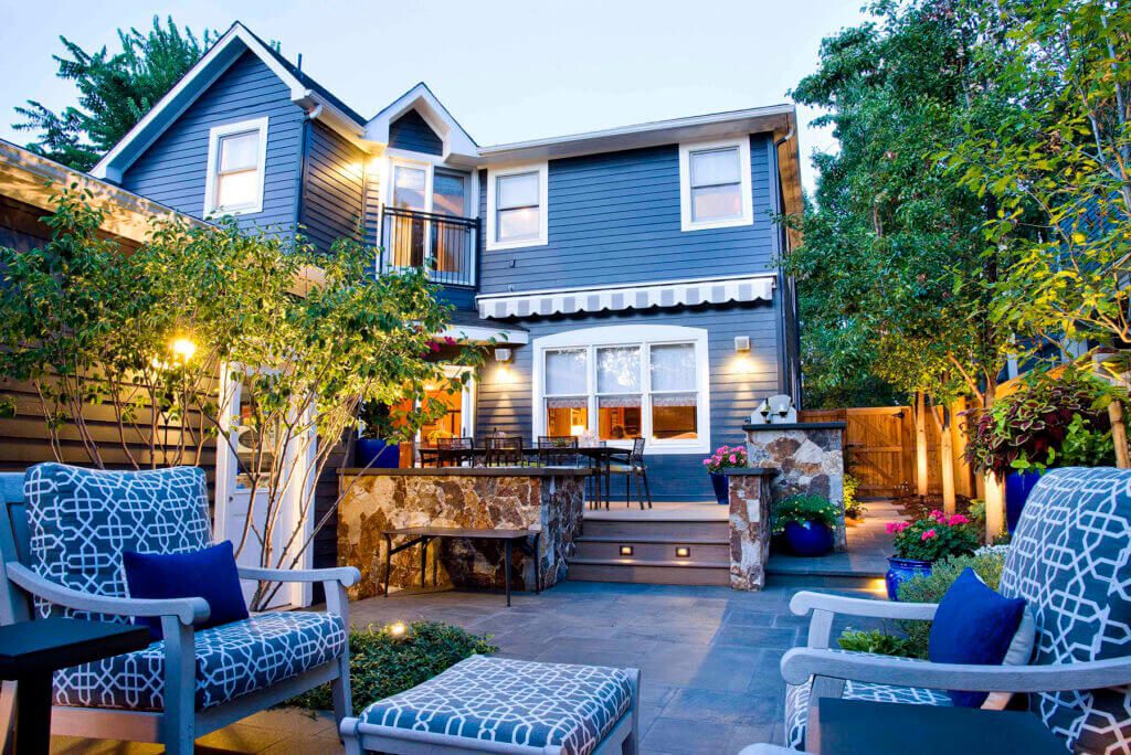 blue house exterior with a stone patio, outdoor seating area with patterned cushions, and a stone-clad raised planting bed with integrated bench seating