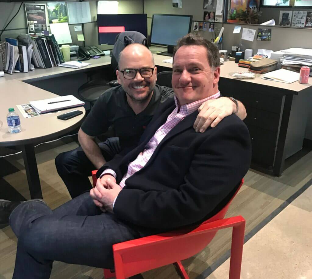 two men smiling and posing together in an office cubicle