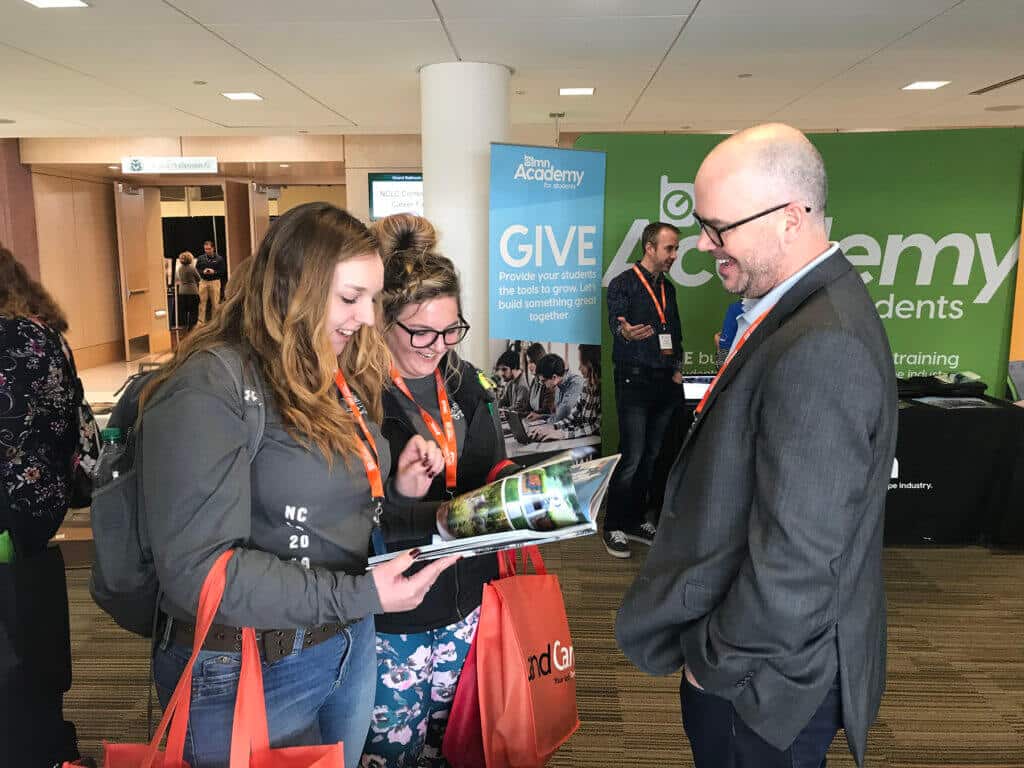 three people at a conference or event looking at a brochure, with a 'Bohme Academy' banner in the background