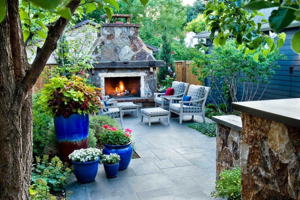 backyard patio with a large stone outdoor fireplace, stylish outdoor seating, and large blue planters with colorful foliage and flowers