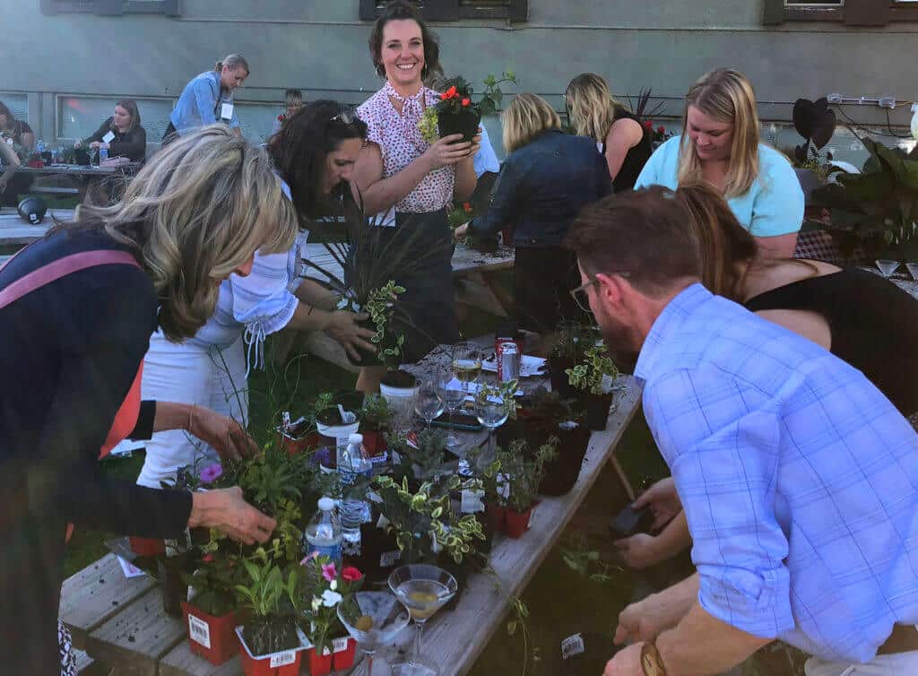 group of people gathered around a wooden table outdoors, planting and potting various plants from small containers
