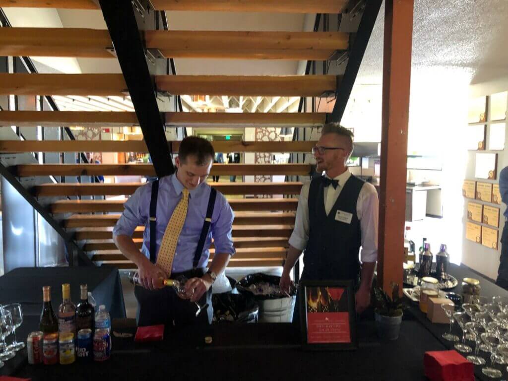 two men working behind a black-draped bar set up indoors beneath an open wooden staircase